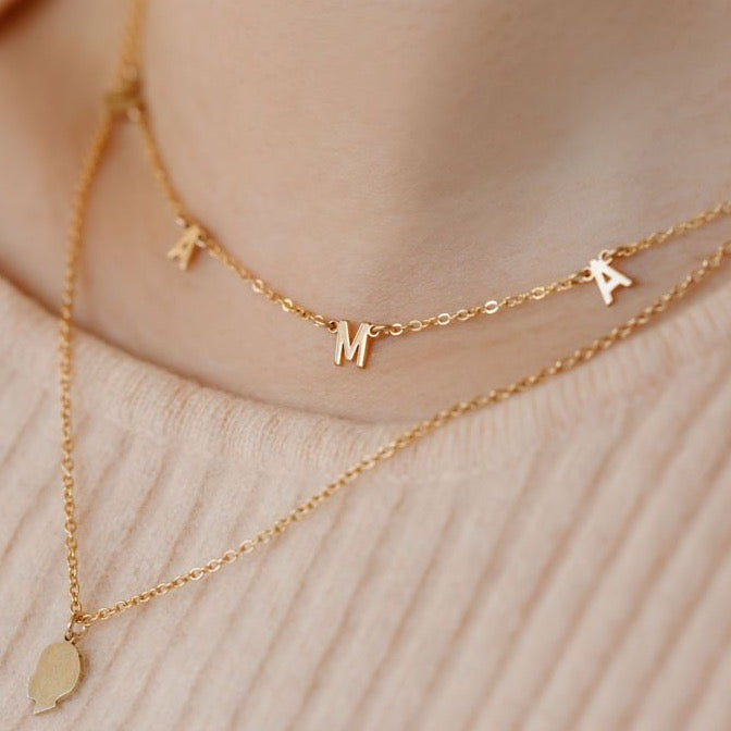 Close up image of layered necklaces, one is a gold kid silhouette necklace 18 inches and the short 16 inch necklace is a gold necklace that spells out MAMA